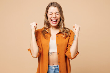 Young Caucasian woman she wears orange shirt casual clothes doing winner gesture celebrate clenching fists say yes close eyes isolated on plain pastel light beige background studio. Lifestyle concept.