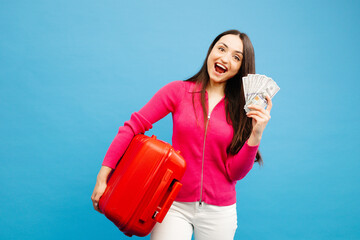 Positive girl with baggage and money are going to travel. Woman in pink sweater and jeans standing on blue background. Vacation concept