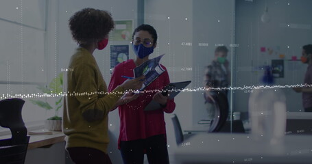 Data processing over two diverse female office colleagues wearing face masks discussing at office