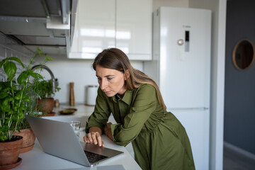 Tired concentrated woman freelancer trying to work at home kitchen standing with laptop after long...