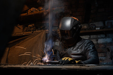 Young mechanic with a welding machine in an old dirty garage at night. Man in protection doing welding work, hobby
