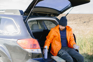 A man rests sitting in the trunk of a car, drinking coffee from a thermos.