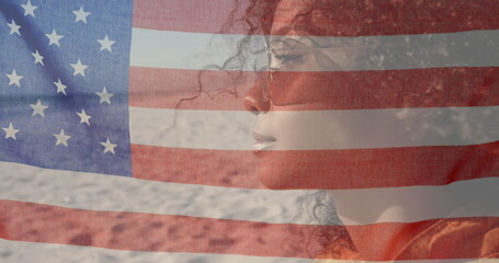 Image of American flag waving over woman with sunglasses sitting on beach by seaside on summer holid