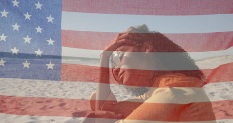 Image of American flag waving over woman with sunglasses sitting on beach by seaside on summer holid