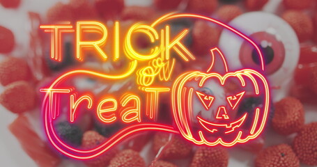 Obraz premium Neon trick or treat text banner with pumpkin icon against scary eye toy and halloween candies