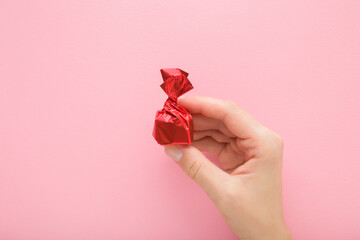 Young adult woman hand fingers holding and showing candy wrapped in bright red foil paper on light pink table background. Pastel color. Closeup. Top down view.