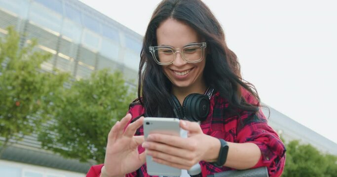 Adorable smiling happy carefree stylish young woman with long brown hair in glasses and with headphones around her neck watching funny videos on her phone during walking on the urban street