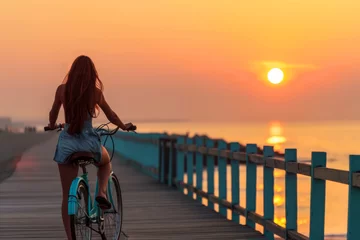 Papier Peint photo Descente vers la plage Silhouette of a woman riding a bike on a beach boardwalk at sunset with ocean view