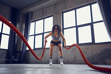 Front view portrait of young athlete woman doing battle rope training creating waves in industrial-style gym. Motivation. Concept of professional sport, bodybuilding, active and healthy lifestyle. Ad