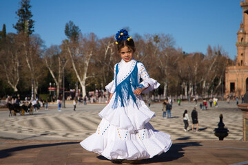 a little girl dancing flamenco dressed in a white dress with ruffles and blue fringes in a famous...