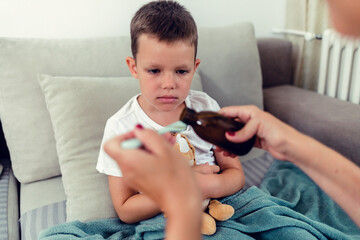 Sick little boy lying in bed making a face at his mother. The mother is giving a spoon of medicine...