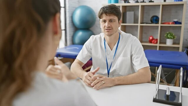 A man in a white coat consults a patient in a well-equipped physiotherapy clinic, featuring rehabilitation balls and a treatment bed.