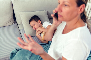 Obraz premium Stressed mother on a call about sick child on a sofa, worried about her son health. Little boy with flu resting, sleeping and waiting for a home visit or checkup by a healthcare professional.