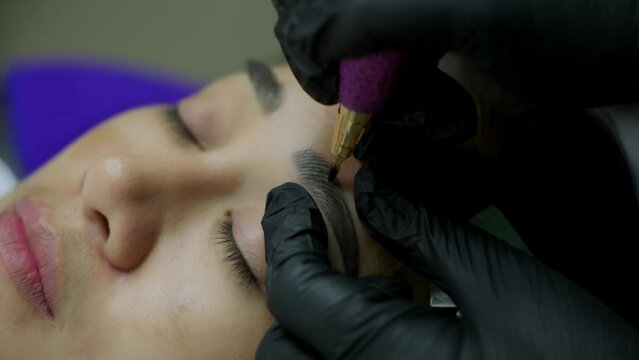 Close up microblading eyebrow tattoo. Beautician in gloves makes permanent makeup correction of the shape of the eyebrows to a young women. Eyebrow tattoo in a beauty salon.