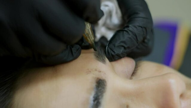 Close up microblading eyebrow tattoo. Beautician in gloves makes permanent makeup correction of the shape of the eyebrows to a young women. Eyebrow tattoo in a beauty salon.