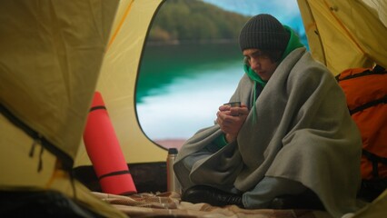 Person on campsite traveling and hiking, exploring nature. Young man covered in blanket drinking hot tea inside the tent at lake coast in mountains, holiday outdoors.