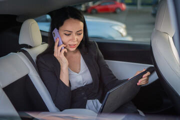 Smiling businesswoman using digital tablet while talking on the smartphone on the back seat of a car. Copy space. Business, road trip, technology, travel concept