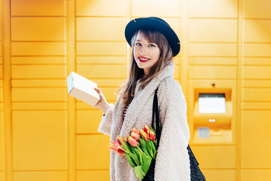 Portrait of smiling fashion woman with box near modern postal automatic mail terminal with self service device for pickup or refund an order. Electronic locker for storing parcels. Selective focus.