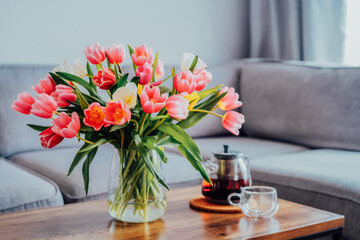 Focus on vase of fresh tulips bouquet and just brewed tea pot and cup on coffee table with gray couch sofa in modern leaving room. Cozy, calm and comfortable relax at home. Time to take a break. - obrazy, fototapety, plakaty