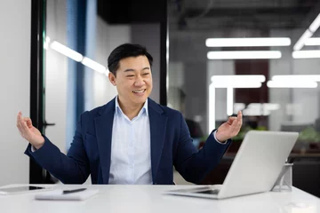 Fotobehang Asian young male businessman sitting at desk in office in lotus position, meditating and looking at laptop screen smiling. He rests and takes a work break. © Liubomir