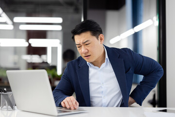 Worried Asian young man sitting in office at desk in front of laptop and holding hand behind back,...