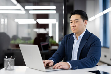 A serious young Asian man in a business suit is sitting in a modern office at a desk and working on a laptop.