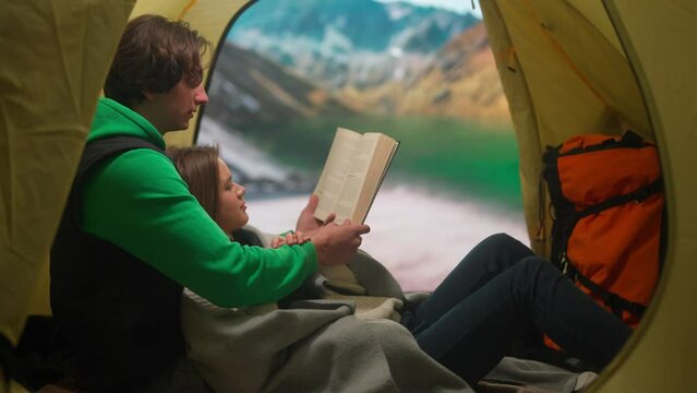 People on campsite traveling and hiking, exploring nature. Young loving couple reading a book inside the tent at lake coast in mountains, holiday and vacation outdoors.
