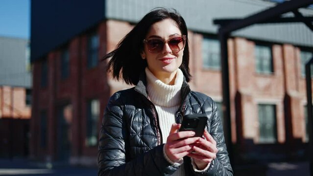 Dark haired woman in sunglasses using new network application on digital phone and smiling at camera. Contemporary tourist using online maps for navigating in city and route planning in real time.