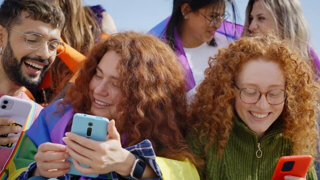 A community of LGBT people sitting using and looking at their mobile phones while sharing pictures with each other, smiling, gesticulating and having fun together outdoor