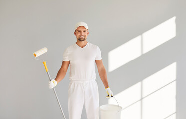 Portrait of a happy smiling male painter in white clothes holding a bucket and paint roller...