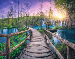 Waterfall and wooden path in green forest. Plitvice Lakes, Croatia at sunset in spring. Colorful landscape with pathway in blooming park, trees, water lilies, river, pink sky in summer. Trail in woods - 758213775