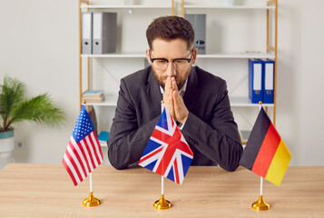 Portrait of a thoughtful pensive young business man in suit sitting at the desk in office with USA, Great Britain and Germany flags choosing country to immigrate or foreign language to study. - obrazy, fototapety, plakaty