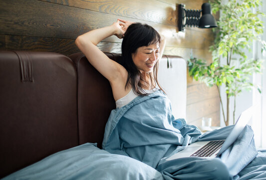 Smiling woman stretching in bed with laptop at home