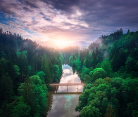 Aerial view of wooden bridge over the river in mountains at foggy summer morning in Slovenia. Top drone view of bridge, green forest, hills, fog, cloudy sky at sunrise in spring. Nature. Road in woods
