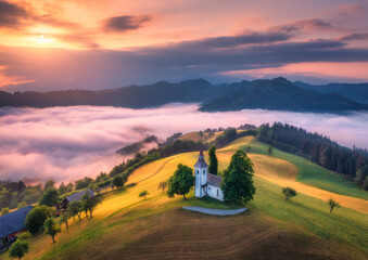 Aerial view of small church on the hill over pink low clouds at foggy sunrise in summer in Slovenia. Top view of beautiful chapel on mountain in fog, green meadows, trees, orange sky at dawn in spring - obrazy, fototapety, plakaty