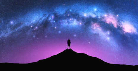 Milky Way arch and man on the mountain peak at starry night. Silhouette of alone guy, pink sky with bright stars in summer. Galaxy. Space background. Landscape with arched milky way. Travel and nature - obrazy, fototapety, plakaty