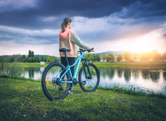 Woman with mountain bike near lake at overcast sunset in spring. Colorful landscape with sporty girl, bicycle, coast of river, green grass, cloudy sky in park in summer. Sport and travel. Biking - 758232763