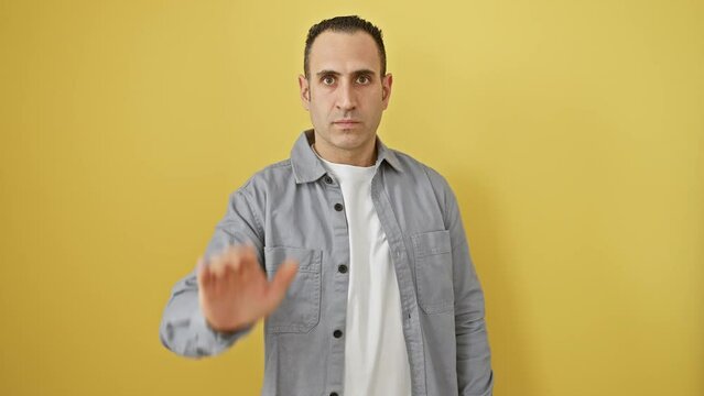 Young hispanic man wearing shirt standing doing stop sing with palm of the hand. warning expression with negative and serious gesture on the face. over isolated yellow background