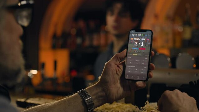 Mature sports fans check game score and bookmaker ratings in app using mobile app phone, talk with bartender. Male friends bet on football match online sitting at bar counter in pub. Gambling concept.