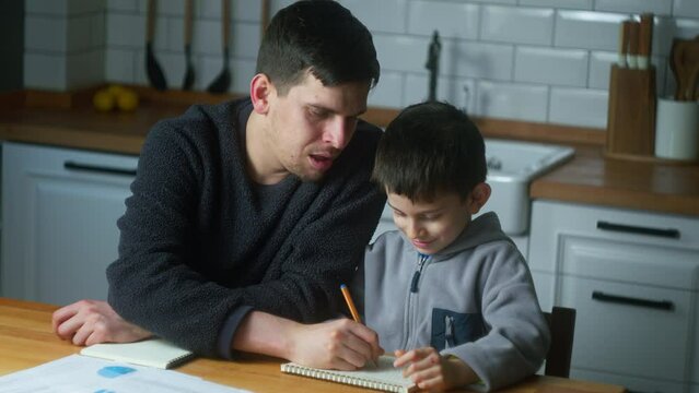 Young father help schoolboy with homework at home. Dad helping to son with writing while sitting in the kitchen. Adult parent or tutor with kid boy learning.