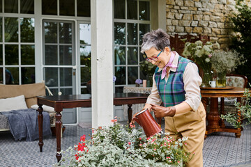 attractive jolly mature woman in chic attire watering her flowers near her house in England