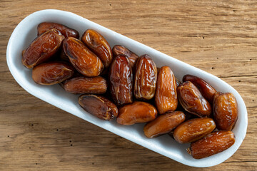 Dry dates fruit in a white container on wooden background, top view, closeup