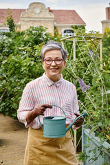 cheerful mature woman with glasses holding watering can and smiling at camera near house in England