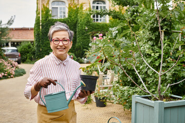 appealing joyful mature woman holding watering can and pot with plant and smiling at camera, England
