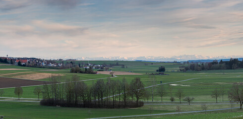 Panorama einer Landschaft mit einem Dorf und Wiesen sowie den Alpen am Horizont