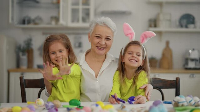 Easter grandmothers with granddaughters. Smiling grandmother hugging twins grandchildren wearing rabbit bunny ears, decorating paint eggs together in kitchen at home. Tradition Easter holiday concept.