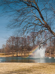Bare trees in a park surround lake with fountain under a blue winter sky