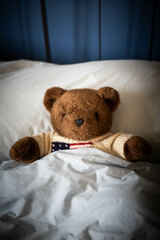 Brown teddy bear, plush toy, laying under covers in parents' bed with blue blankets and soft day light. Central view, vertical shot.