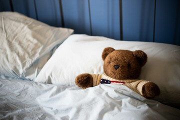 Brown teddy bear, plush toy, laying under covers in parents' bed with blue blankets and soft day light. Side view, background with copy space..