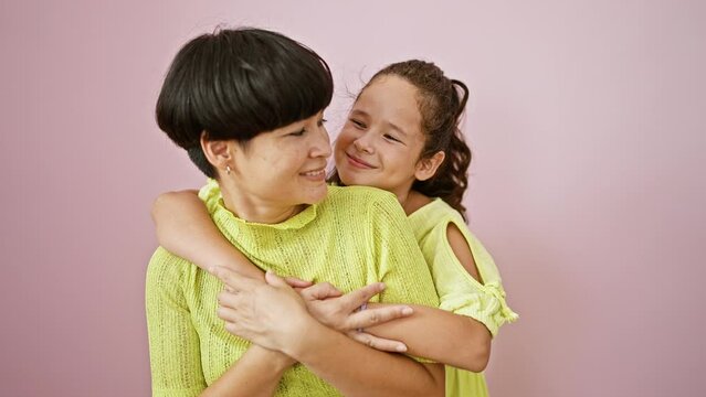 Confident mother and daughter sharing a lovely, happy hug while smiling and standing together over an isolated pink background, expressing a casual lifestyle full of fun, joy and positive vibes.